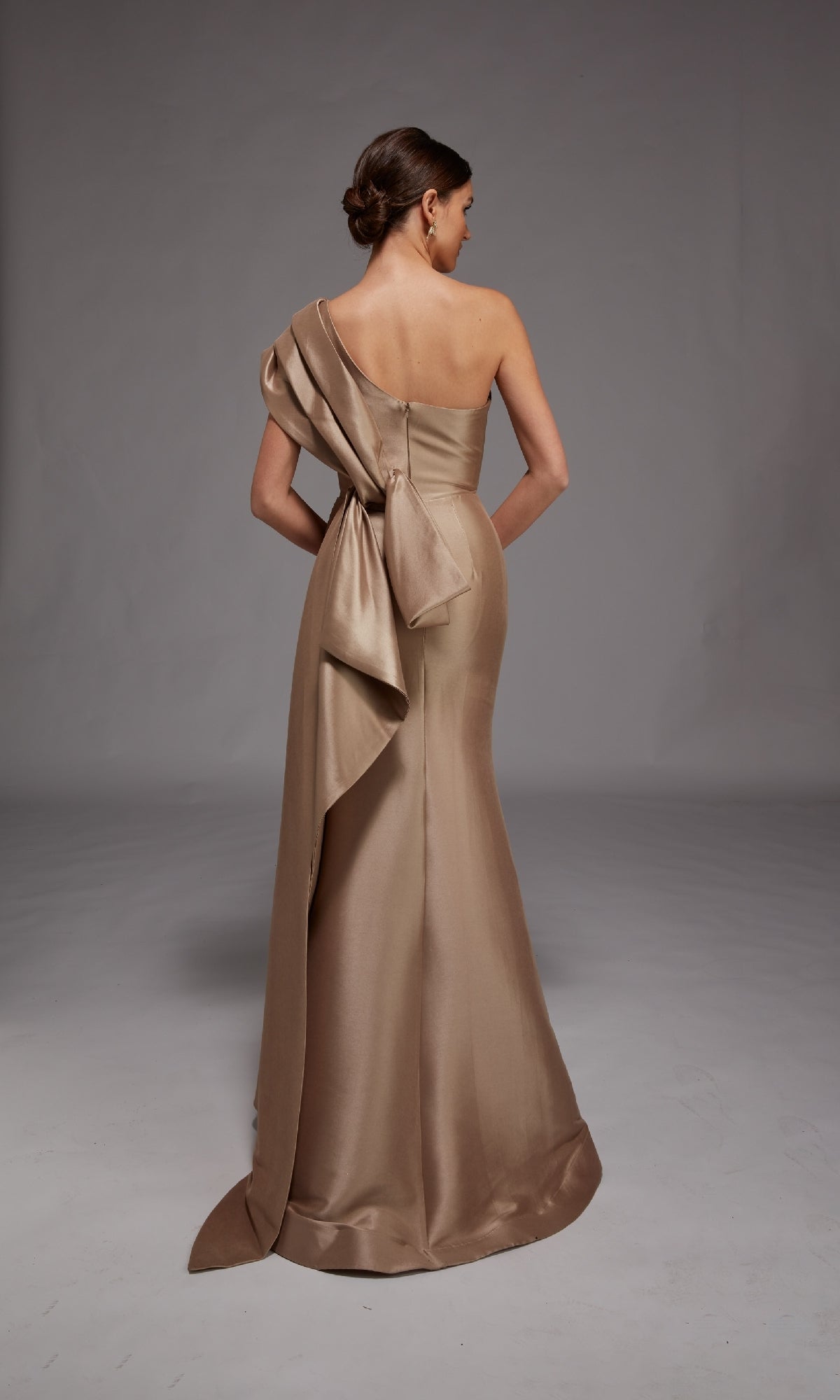 A woman stands against a plain gray background, wearing the Alyce 27838—a long formal satin gown featuring a one-shoulder design, floor-length silhouette, and a striking large bow at the back.